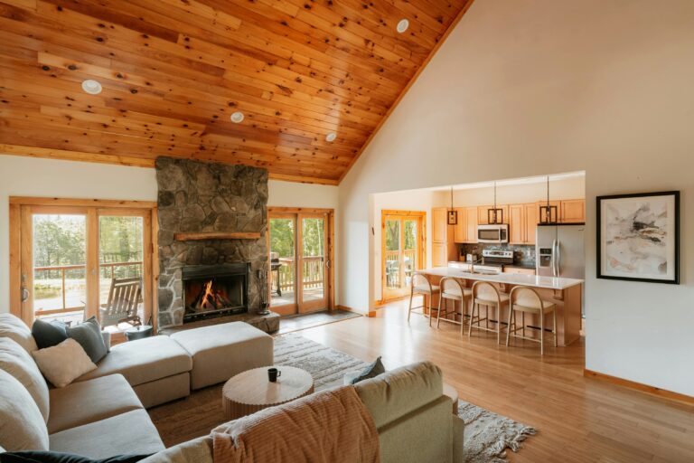 Open concept remodel in a cabin with a vaulted pine ceiling, a large stone fireplace, and a light-filled living area connected to a modern kitchen with natural wood cabinets and a breakfast bar.
