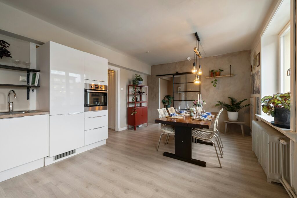 Open concept remodel showing a modern kitchen with sleek white cabinets flowing into a warm dining area with a wooden table, hanging lights, and laminate flooring.