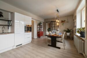 Open concept remodel showing a modern kitchen with sleek white cabinets flowing into a warm dining area with a wooden table, hanging lights, and laminate flooring.