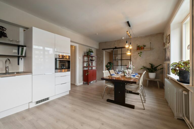 Open concept remodel showing a modern kitchen with sleek white cabinets flowing into a warm dining area with a wooden table, hanging lights, and laminate flooring.